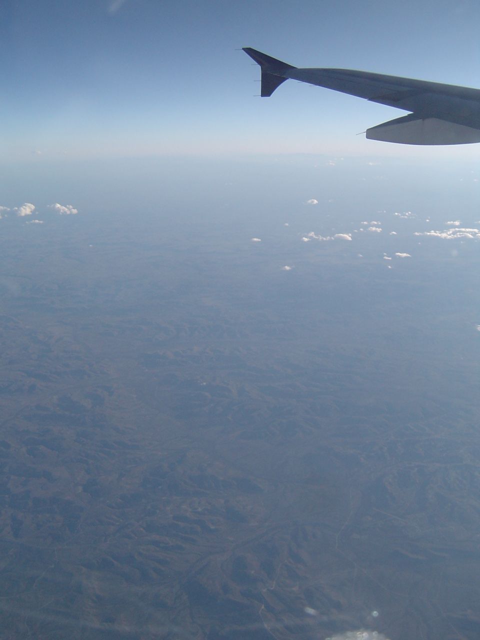 Airplane wing against clear blue sky, with hazy landscape below
