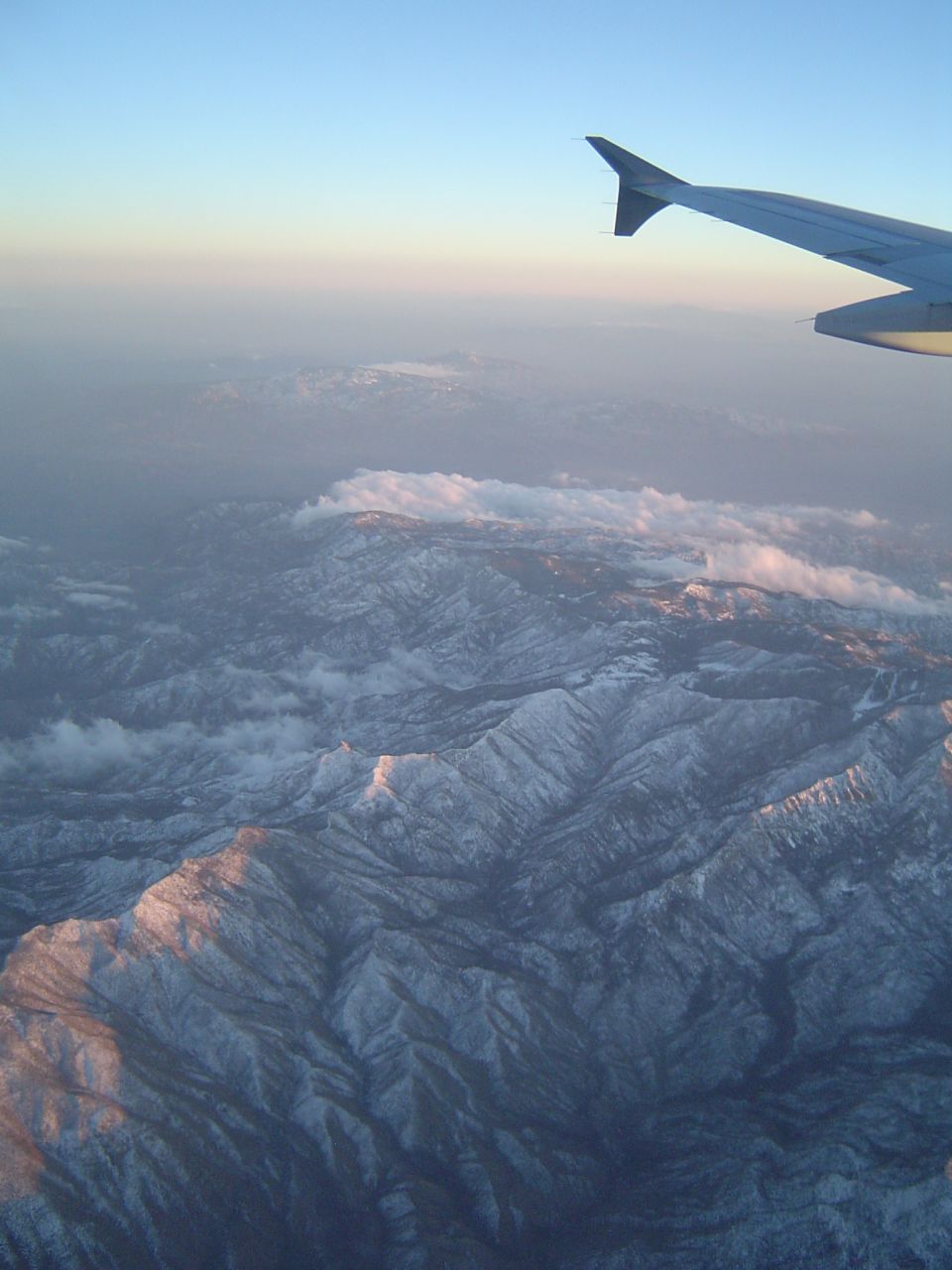 Airplane wing in upper right corner, overlooking vast snow-covered mountains at sunrise