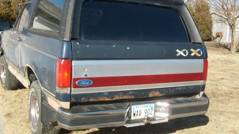 1995 Ford C-150 (Centurion Conversions) - Rear view of a blue Ford Bronco with a silver and red stripe on the tailgate, showing significant rust.
