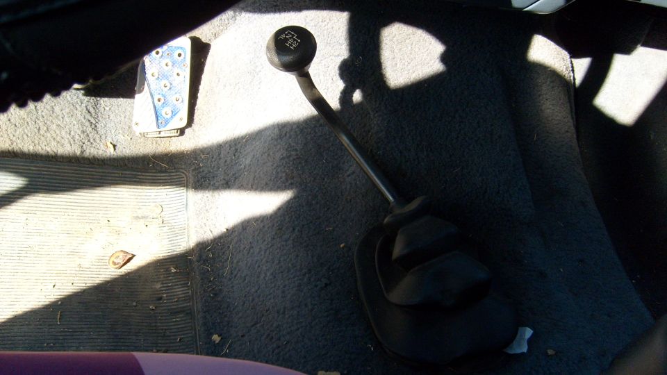 Black five-speed manual shifter and boot on gray carpet with blue drilled gas pedal