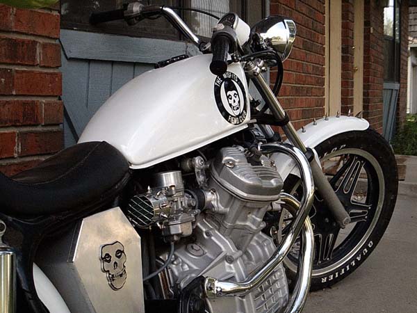 White custom bobber motorcycle with skull logo tank and twin engine parked by brick wall