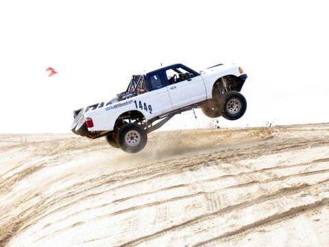 1984 Ford Bronco - A white off-road truck is captured mid-air as it jumps over a sand dune.