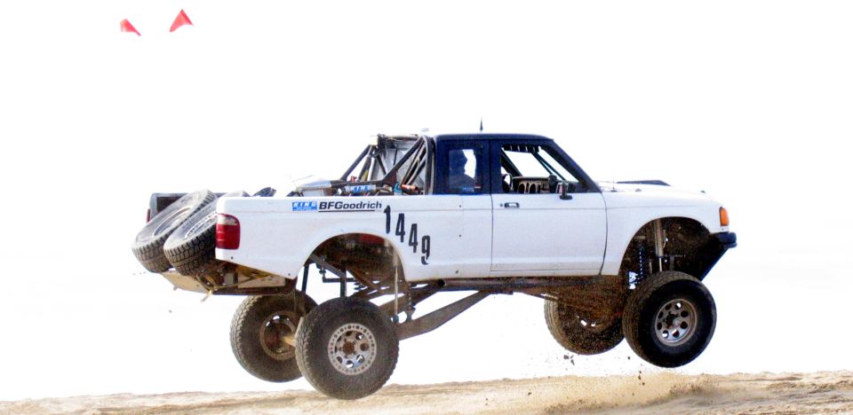 White Ford Ranger off-road truck with roll cage, spare tires on rear, jumping. BFGoodrich tires visible