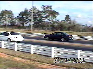 1997 Ford Taurus - Two sedans are racing down a drag strip on a sunny day, with trees and a fence in the background.