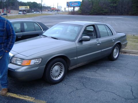 1996 Ford Crown Victoria - A gray sedan is parked in a lot next to another car, with a billboard visible in the background.