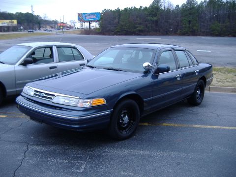 1996 Ford Crown Victoria - Two sedans, a dark blue 1996 Ford Crown Victoria and a silver one, are parked in an asphalt lot.