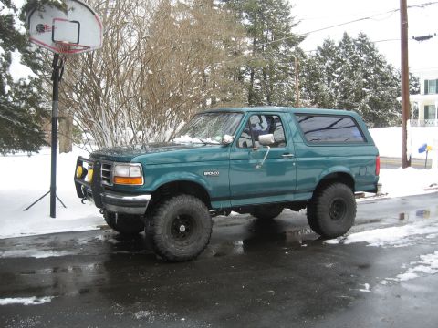 1996 Ford Bronco - A green 1996 Ford Bronco sits on a wet road with snow covering the surrounding trees and ground.