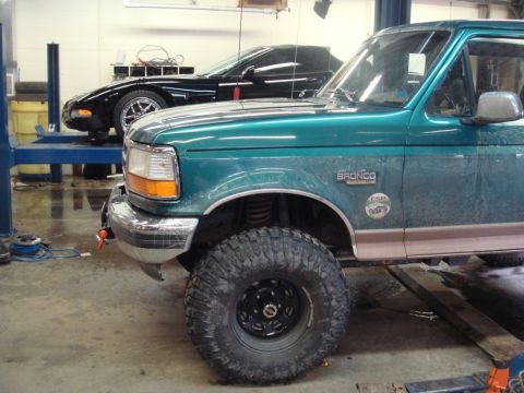 1996 Ford Bronco - A green 1996 Ford Bronco with prominent off-road tires is shown in a garage, with a black sports car in the background.