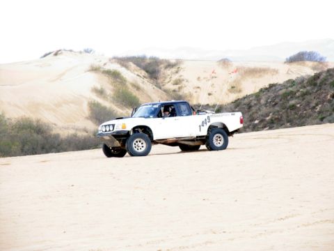 1984 Ford Bronco - Modified pickup truck navigates sandy terrain with dunes in the background.