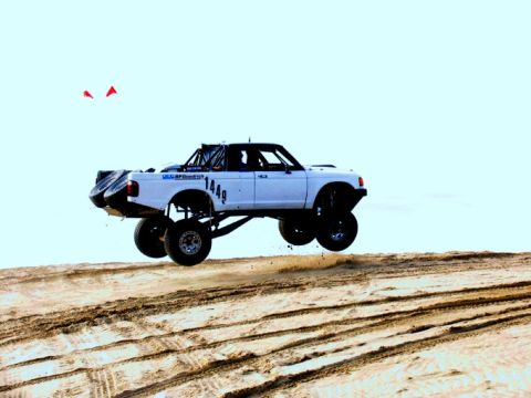 1984 Ford Bronco - A modified pickup truck is captured mid-air, soaring over a sandy terrain under a clear sky.