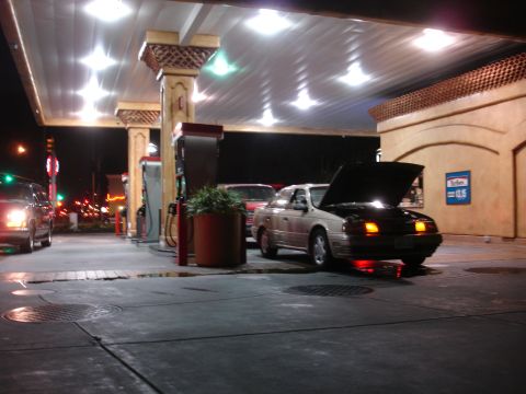 1991 Ford Taurus - A beige 1991 Ford Taurus sits at a gas pump with its hood open at night.
