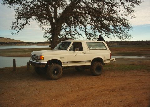 1987 Ford Bronco - This lifted Ford Bronco is parked near a body of water with a large tree overhead.