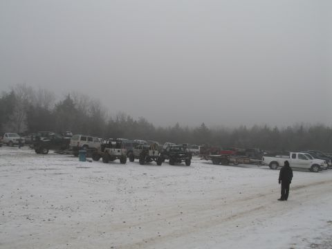 1995 Ford Bronco - A variety of vehicles are parked in a snowy lot, with a person standing in the foreground and trees in the background.