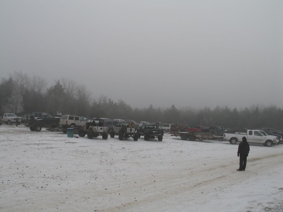 Snow-covered parking lot with many trucks and SUVs, some on trailers, with a treeline in the background and a person standing in the foreground
