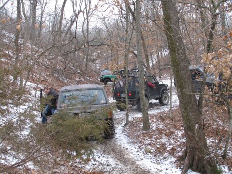 1995 Ford Bronco - Multiple off-road vehicles navigate a challenging, snow-dusted trail in a dense forest.