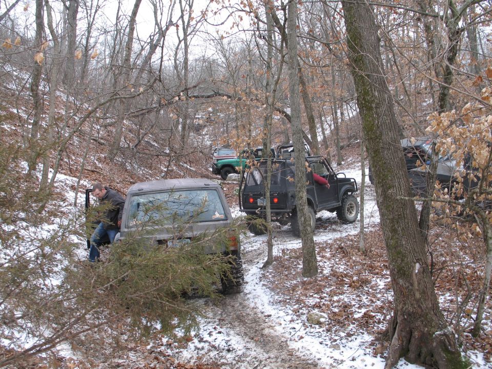 Several off-road vehicles driving on a muddy, snowy trail through a wooded area