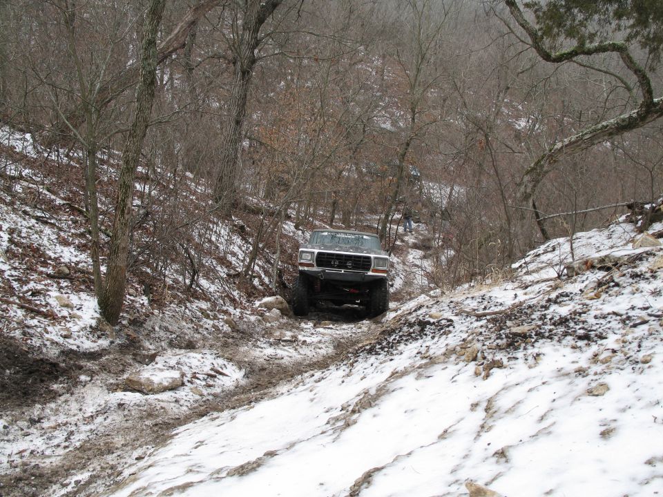 1995 Ford Bronco navigating a steep, snowy, and muddy off-road trail with trees on either side
