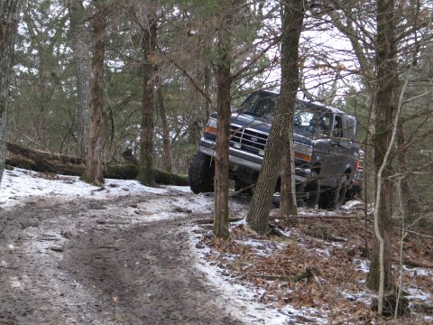 1995 Ford Bronco - A dark 1995 Ford Bronco navigates a challenging muddy and snowy path through a dense forest.