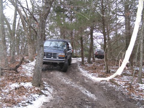 1995 Ford Bronco - Two SUVs navigate a muddy and snowy forest trail, suggesting off-road activity.