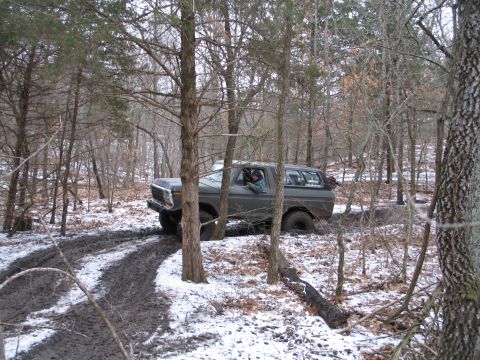 1995 Ford Bronco - A gray SUV navigates a muddy and snowy trail through a forest.