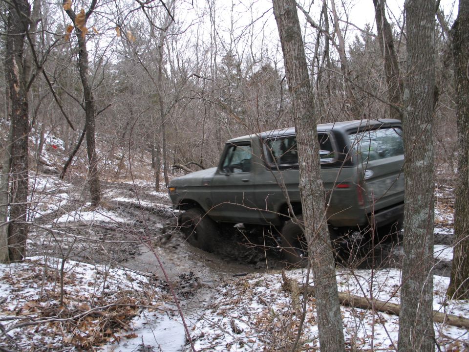 Olive green Ford Bronco driving through a muddy, wooded area with snow on the ground