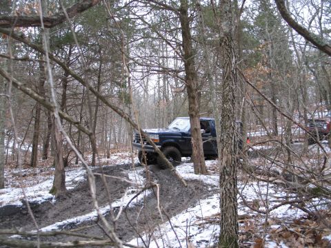 1995 Ford Bronco - A dark blue SUV navigates a muddy, snow-dusted trail through a wooded area.