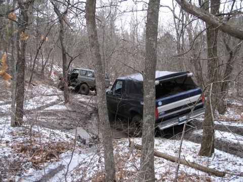 1995 Ford Bronco - Two SUVs are off-roading on a muddy, snow-dusted trail through a forest.