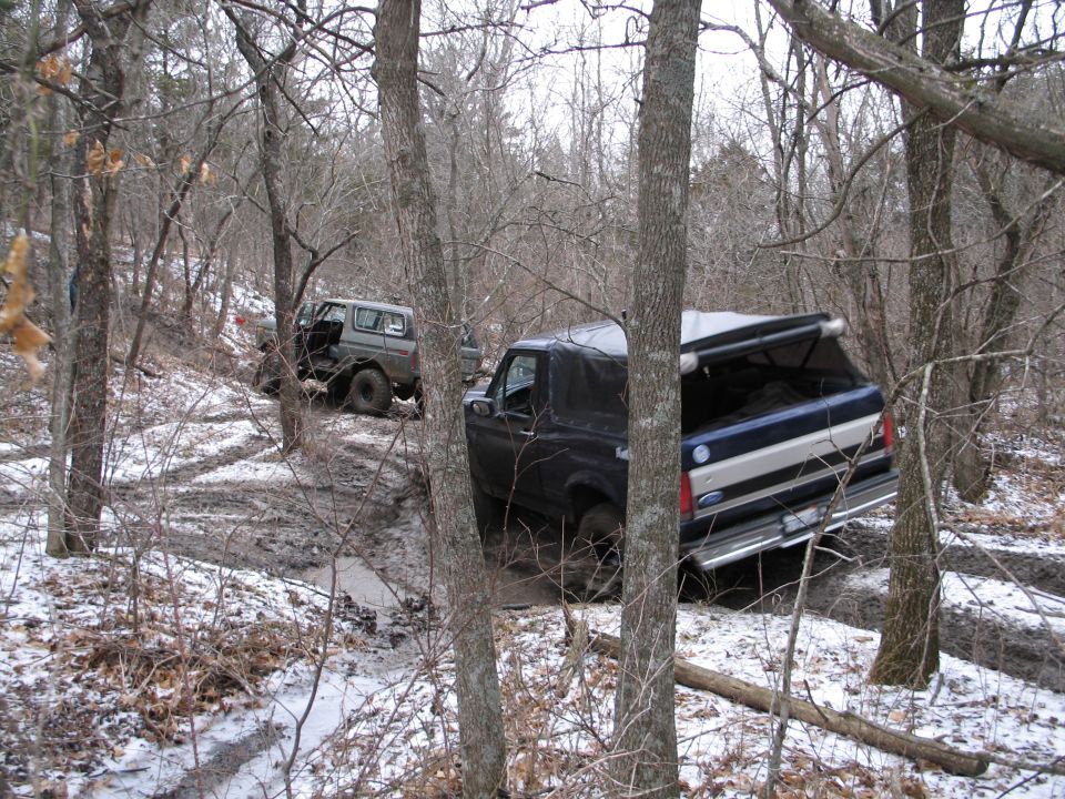 Two SUVs, one dark blue and one grey, are on a muddy trail in a wooded area with snow on the ground. The blue SUV has its rear hatch open