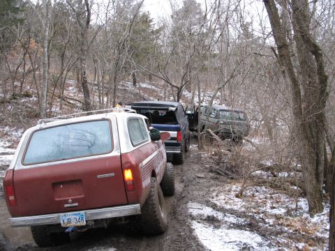 1995 Ford Bronco - A line of SUVs and trucks navigate a challenging, muddy path in a forest during winter.