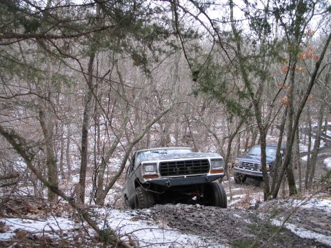 1995 Ford Bronco - Two off-road vehicles navigate a muddy and snowy trail through a wooded area.