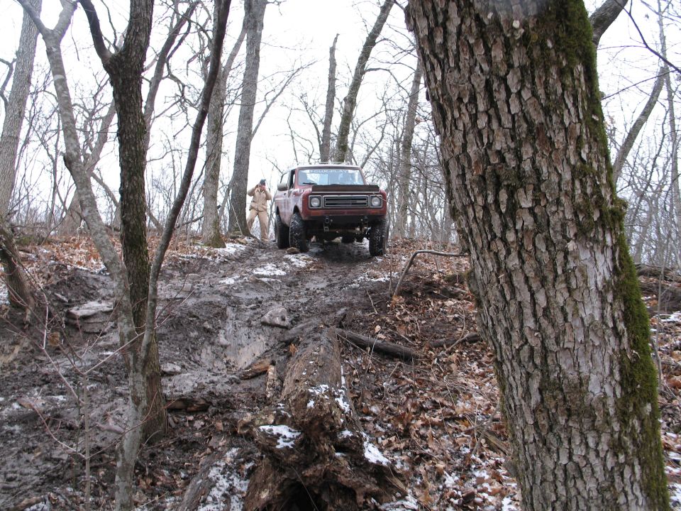 Red off-road truck navigating a steep, muddy incline in a wooded area with bare trees