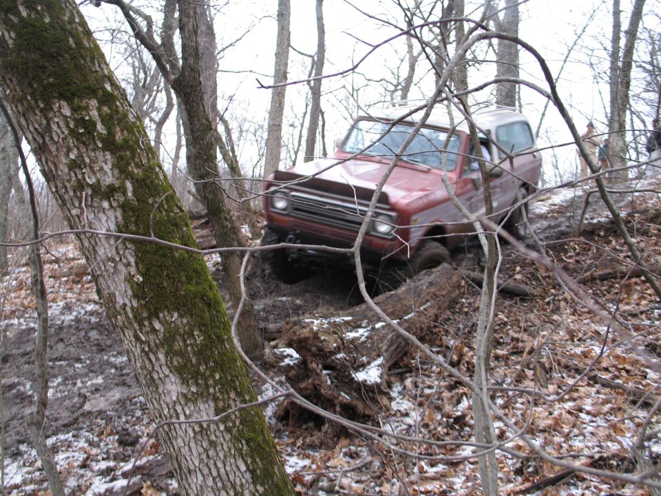 Red SUV with a white top navigating a muddy, wooded trail over fallen logs