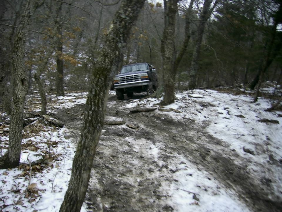 Dark blue Ford Bronco driving on a muddy, snowy trail through trees