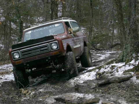 1978 Ford Bronco - An off-road vehicle navigates a challenging, wooded trail with patches of snow and mud.