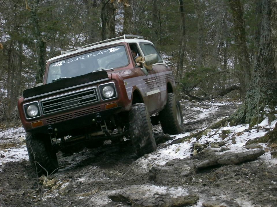 Red International Harvester Scout II off-roading on a muddy, snowy trail in the woods. Windshield sticker says "FOUR PLAY"