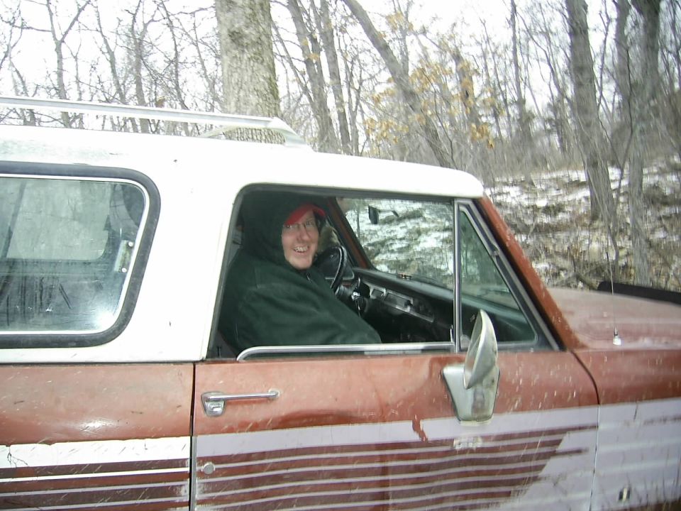 Person wearing a red hat and green jacket inside a rusty SUV with a white roof and rack. Trees and snow in background