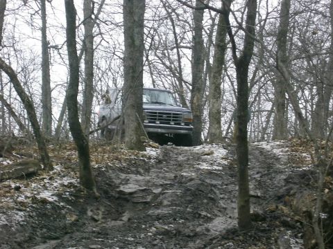 1978 Ford Bronco - A dark SUV navigates a challenging muddy path through a bare, winter forest.