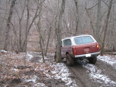 1995 Ford Bronco - A red and white SUV navigates a muddy, wooded trail with patches of snow on the ground.