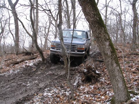 1995 Ford Bronco - A Ford Bronco navigates a challenging muddy path through a bare, wooded landscape.