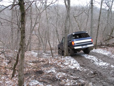 1995 Ford Bronco - A blue Ford Bronco navigates a challenging muddy and snowy off-road trail surrounded by bare trees.