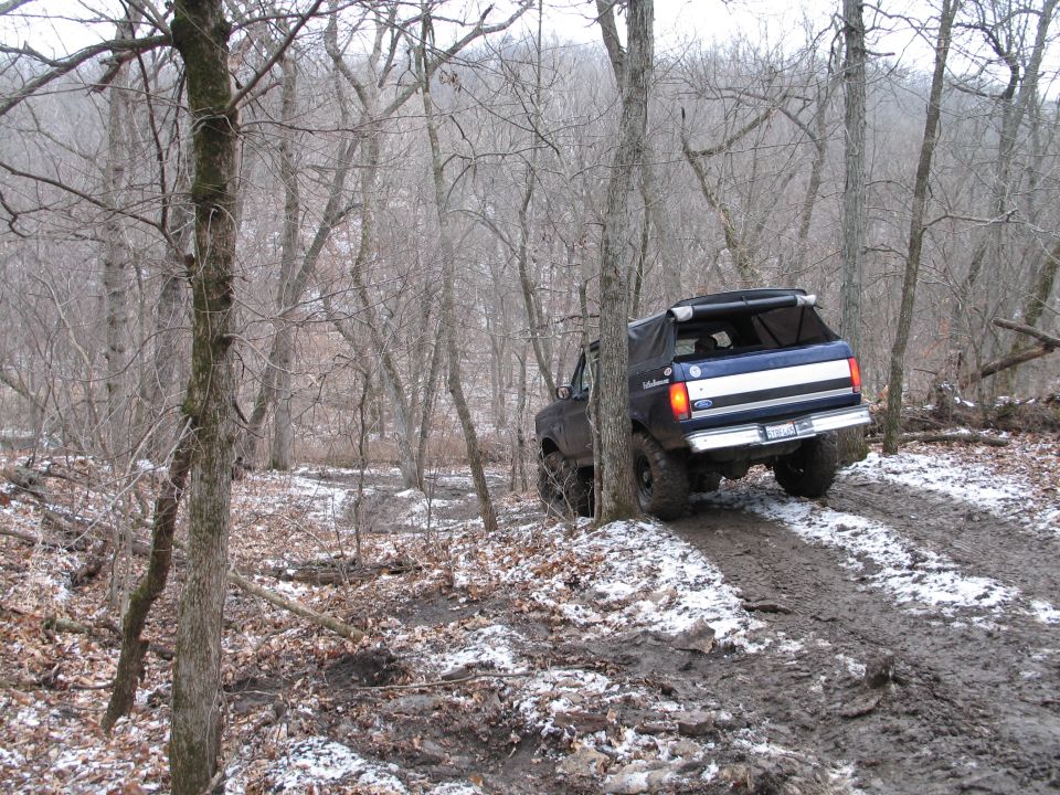 Blue Ford Bronco with a white stripe on the rear, driving on a muddy, snowy trail through trees