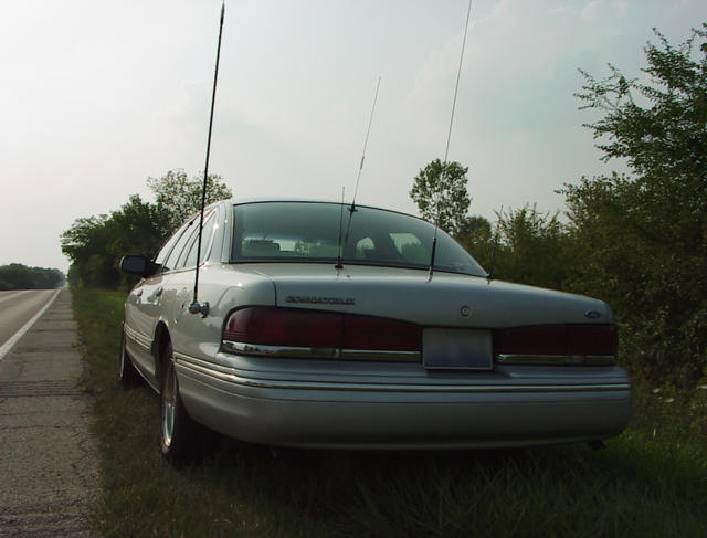 Silver sedan parked on the side of a road, rear view. It has multiple antennas and appears to be from the 1990s