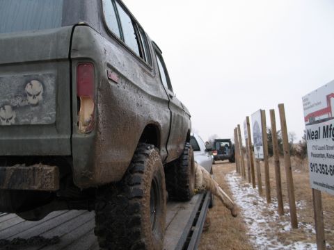 1995 Ford Bronco - A heavily muddied SUV is loaded onto a trailer, with another vehicle visible in the background and signage along the roadside.