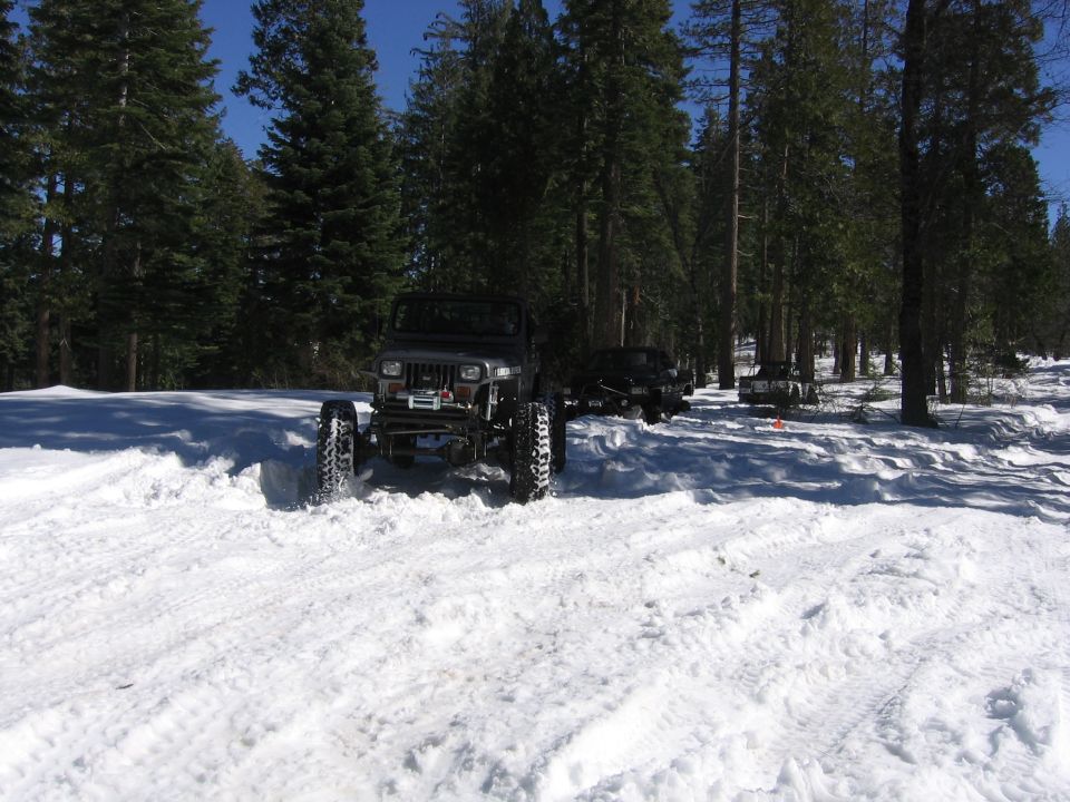 Jeep Wrangler driving through deep snow in a forest with other vehicles behind it