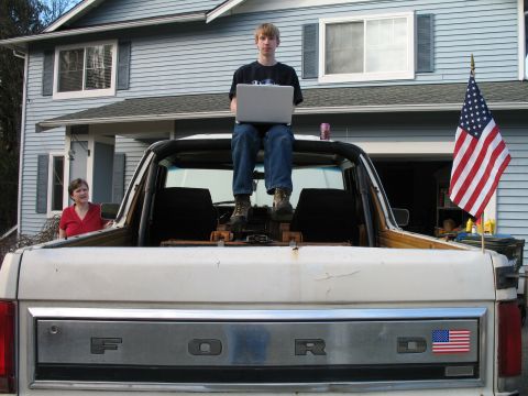 1980 Ford Bronco - A person sits on the tailgate of a Ford pickup truck with a laptop, an American flag flies from the fender.