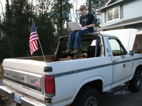 1980 Ford Bronco - A white 1980 Ford Bronco is seen from the rear, with an American flag flying and a person using a laptop on top.