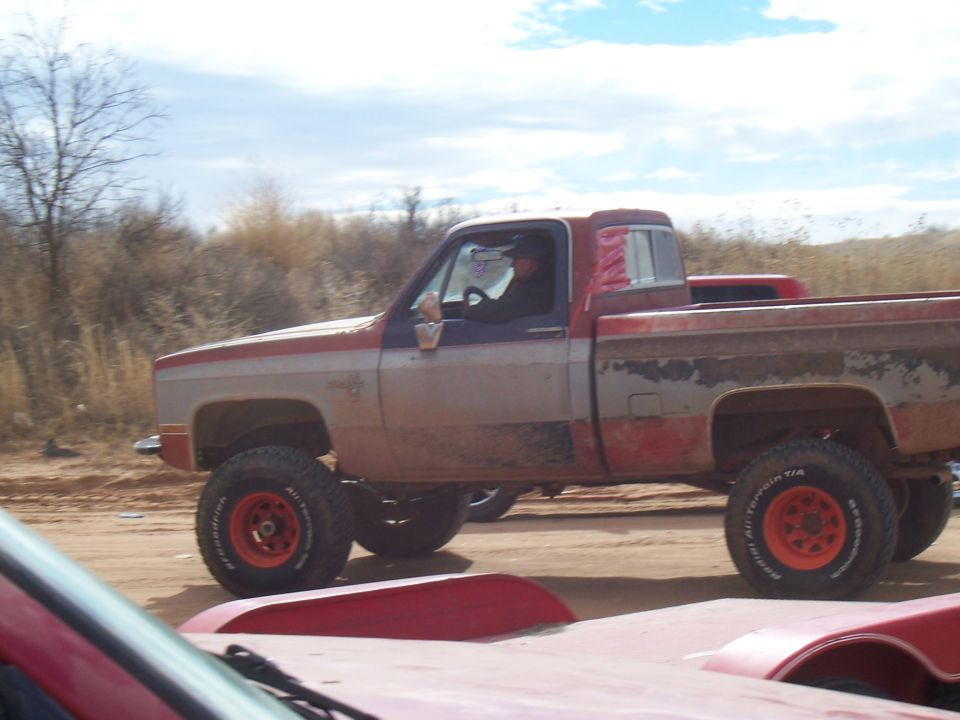 Lifted red and gray pickup truck driving on a dirt road. Another red vehicle is in the foreground