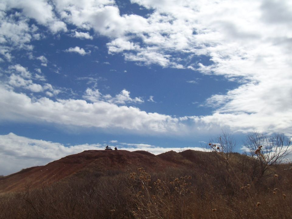 Two ATVs parked on a red dirt hill under a blue sky with white clouds