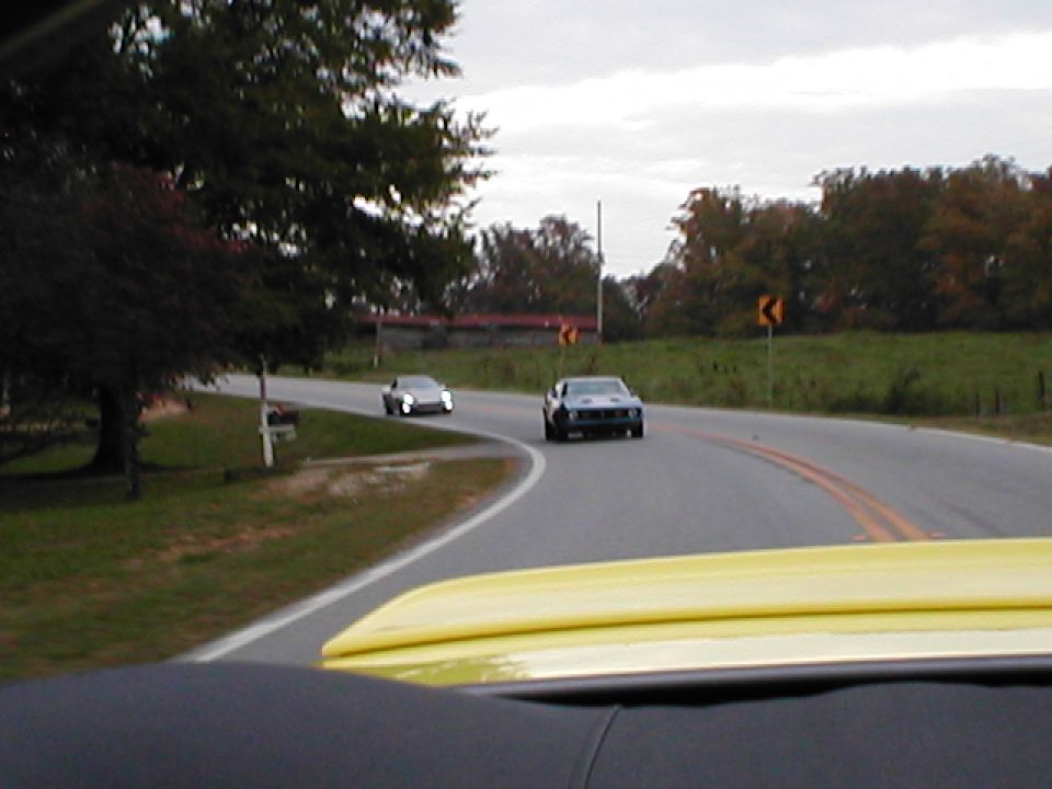 Dark gray 1970s era muscle car driving on a two-lane road, with a silver sedan behind it