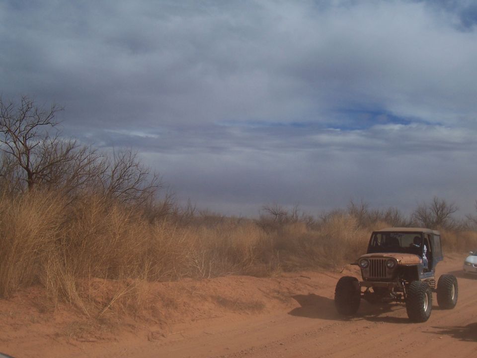 Brown off-road vehicle with large tires on a dusty dirt road, another white vehicle behind it